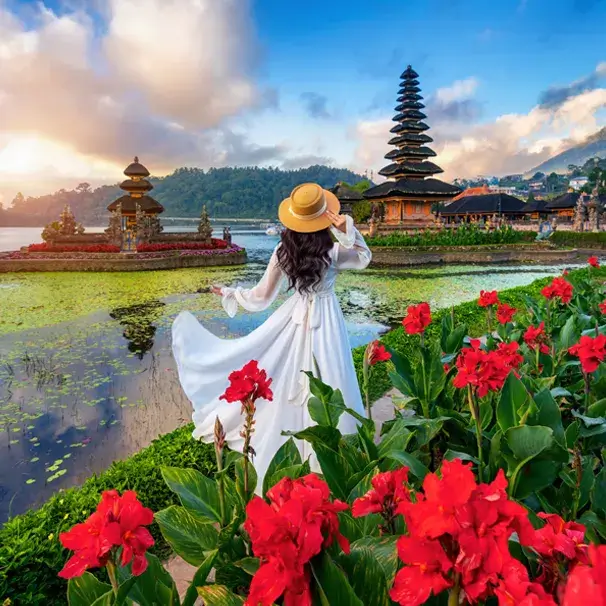 Woman in a white dress and hat gazes at a serene lake surrounded by vibrant flowers in Bali, Indonesia.