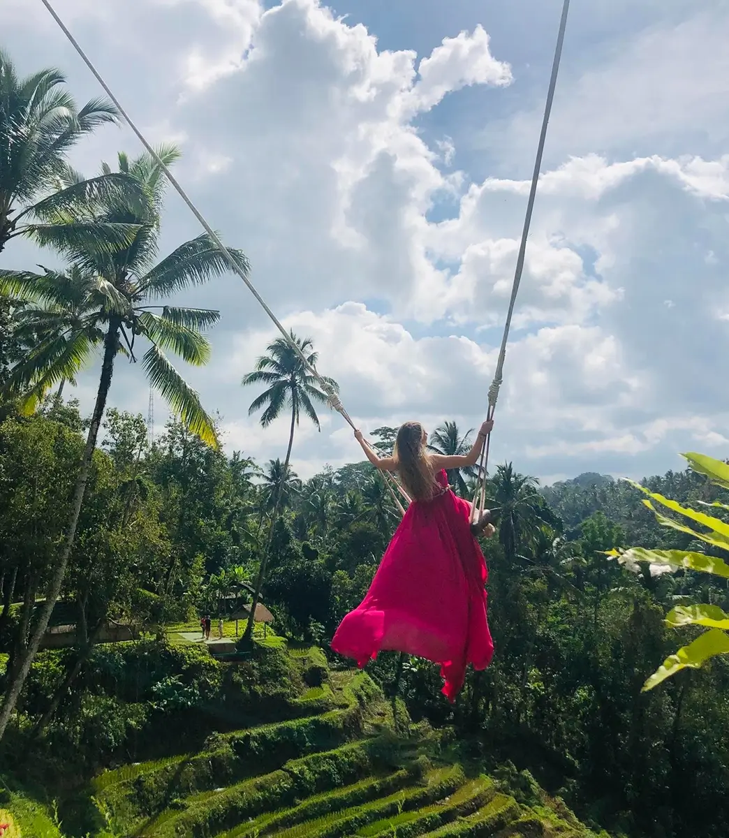 Woman in a flowing pink dress is on a high swing, soaring over lush green terraced rice fields and palm trees under a cloudy sky