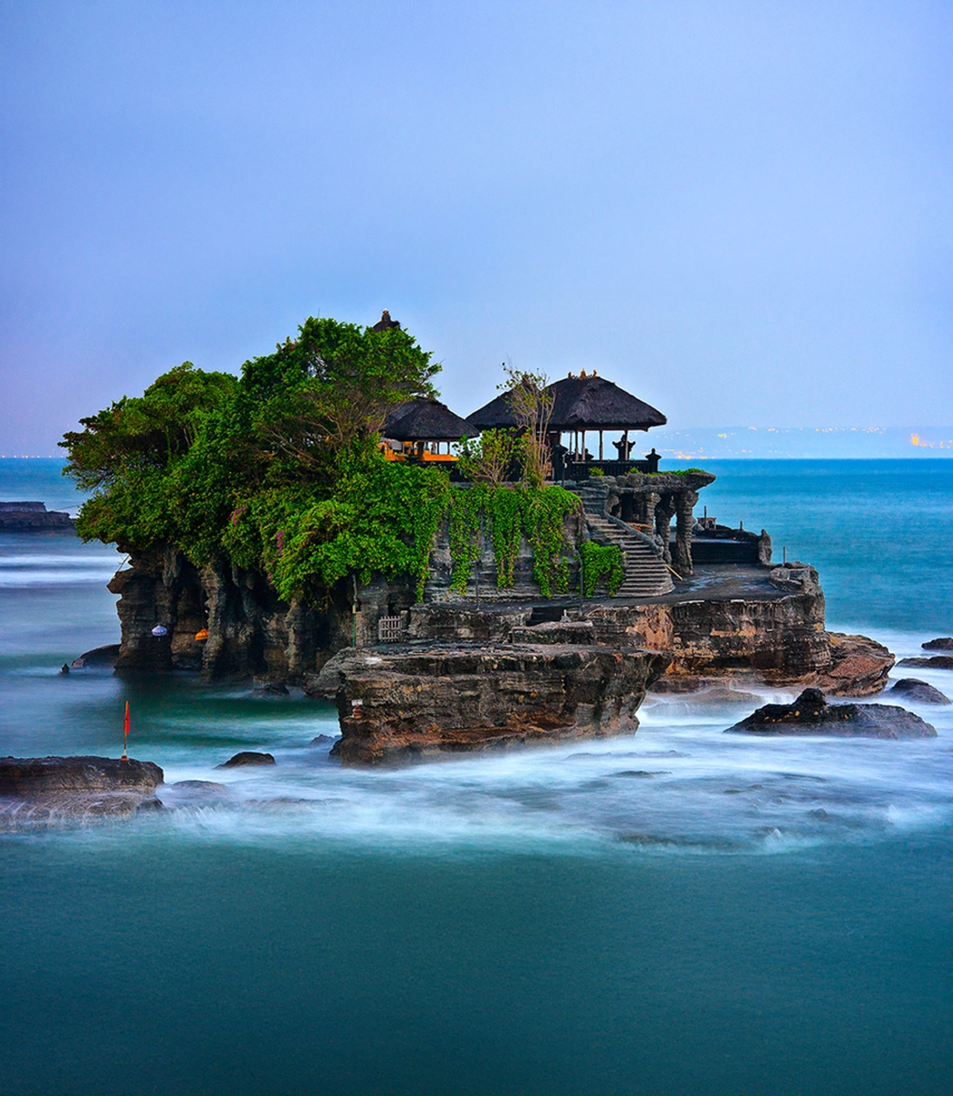 Tanah Lot Temple, a traditional Balinese temple with pagoda-style roofs and lush green trees, sits atop a rocky island surrounded by milky, blurred blue ocean water under a twilight sky.
