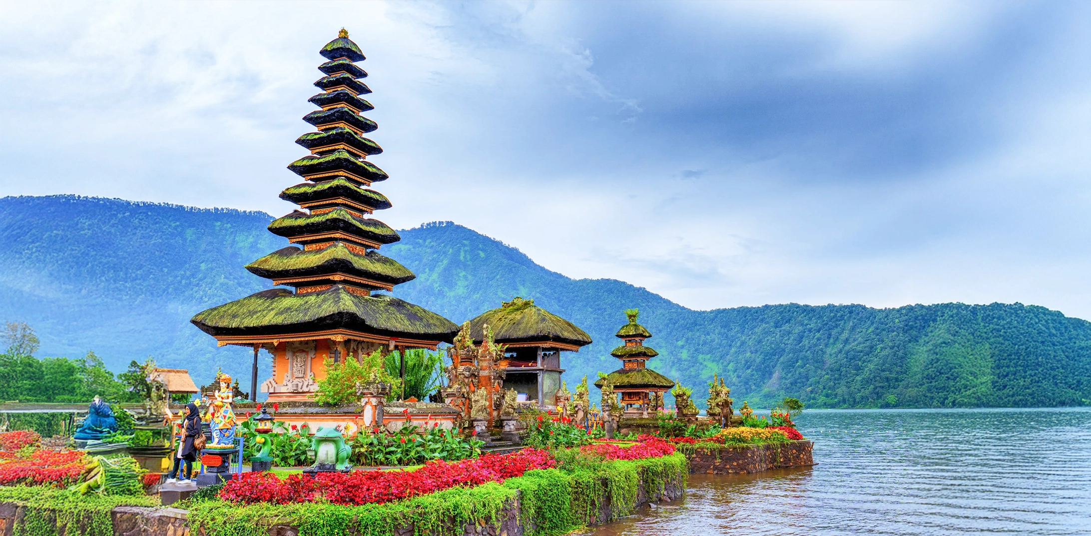 A multi-tiered pagoda-style temple stands beautifully on the water's edge in Bali, Indonesia, surrounded by lush gardens and mountains.