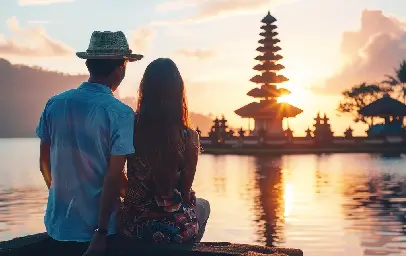 A couple sits with their backs to the viewer, watching a vibrant sunset over a lake with a traditional multi-tiered Balinese temple silhouetted in the distance against the golden sky.