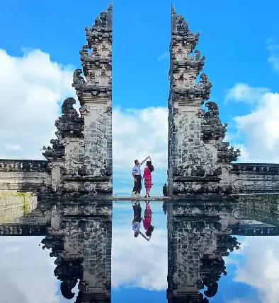 Couple posing between the iconic split gates of Lempuyang Temple in Bali, with a perfect reflection in the water below and a blue sky.