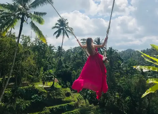 Woman in a flowing pink dress is on a high swing, soaring over lush green terraced rice fields and palm trees under a cloudy sky