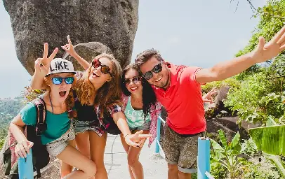 A group of six friends gathered on a bali beach during sunset, toasting with drinks around a barbecue grill, with a palm tree and thatched hut in the background.