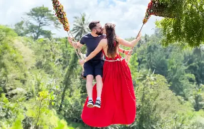 Honeymooners in Bali embrace under a romantic beachfront canopy draped with white and blue fabric and decorated with flowers and palm fronds, beside a candlelit table, with tiki torches, sailboats, and a vibrant sunset over the ocean in the background.