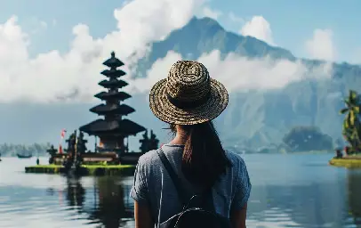 Woman wearing a straw hat and backpack stands with their back to the viewer, looking out at a multi-tiered Balinese temple on a lake, with a large mountain range and cloudy sky in the background.