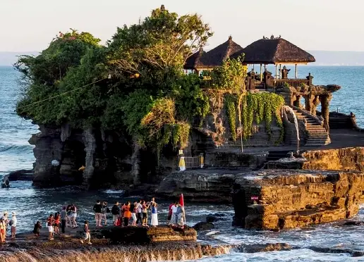 Tanah Lot Temple, a traditional Balinese temple with pagoda-style roofs and lush green trees, sits atop a rocky island surrounded by milky, blurred blue ocean water under a twilight sky.