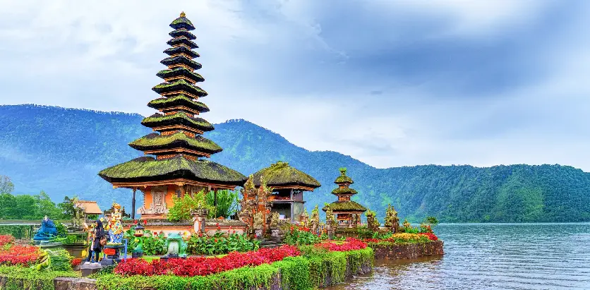 A multi-tiered pagoda-style temple stands beautifully on the water's edge in Bali, Indonesia, surrounded by lush gardens and mountains.