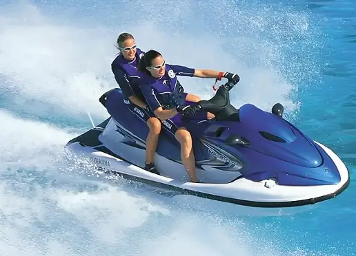 Two people on a blue and white Yamaha jet ski speed across clear blue water, creating a large white spray, both wearing dark wetsuits, sunglasses, and life vests.