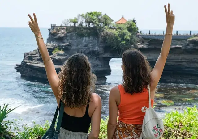 Two women joyfully raise their hands in front of the ocean during their Bali vacation, embracing the sunny beach atmosphere.