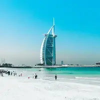 A sunny beach scene with people strolling, featuring the iconic Burj Al Arab hotel in the background.