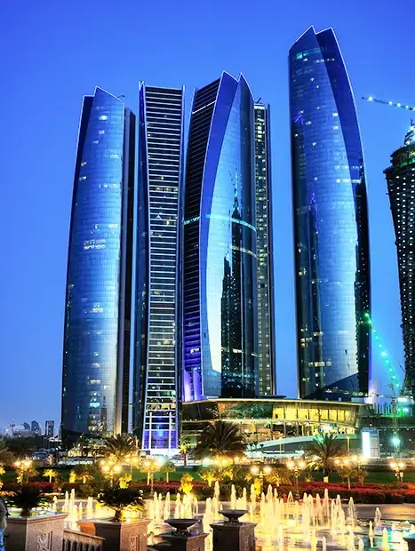 The glowing blue glass facades of the Etihad Towers in Abu Dhabi at dusk with illuminated fountains.