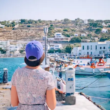 A woman in a hat gazes at boats in a picturesque harbor, capturing the essence of Greece's stunning coastal beauty.