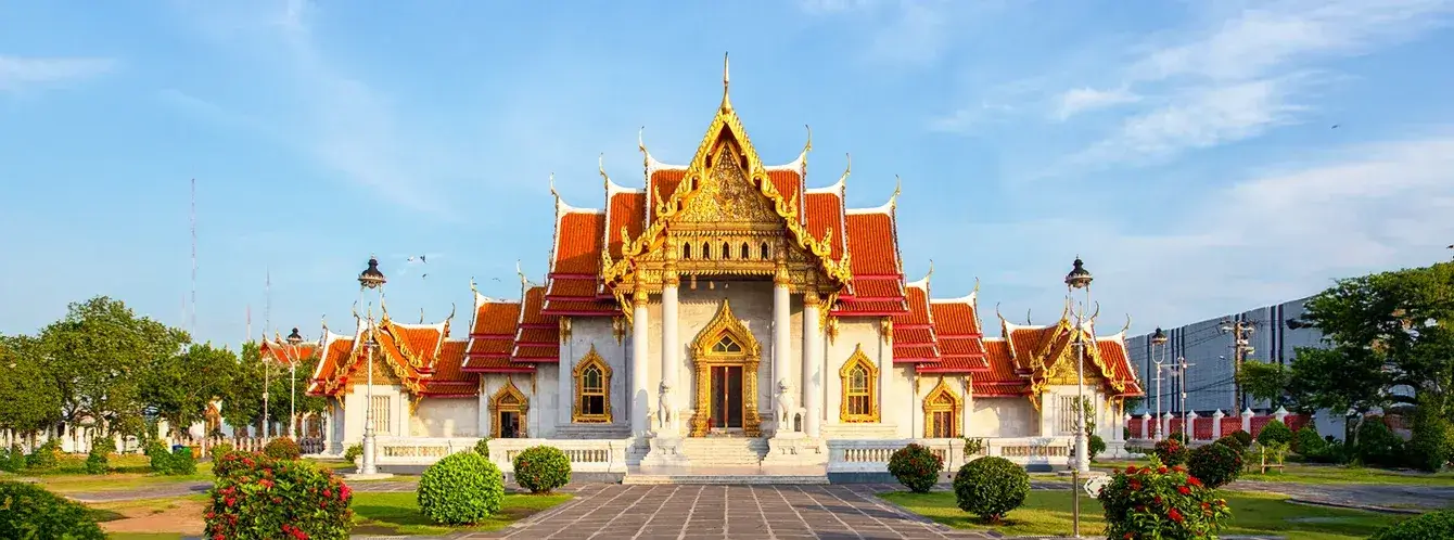 Traditional Thai temple in Bangkok with golden architectural details, red roofs, landscaped gardens, and clear blue sky in daylight.